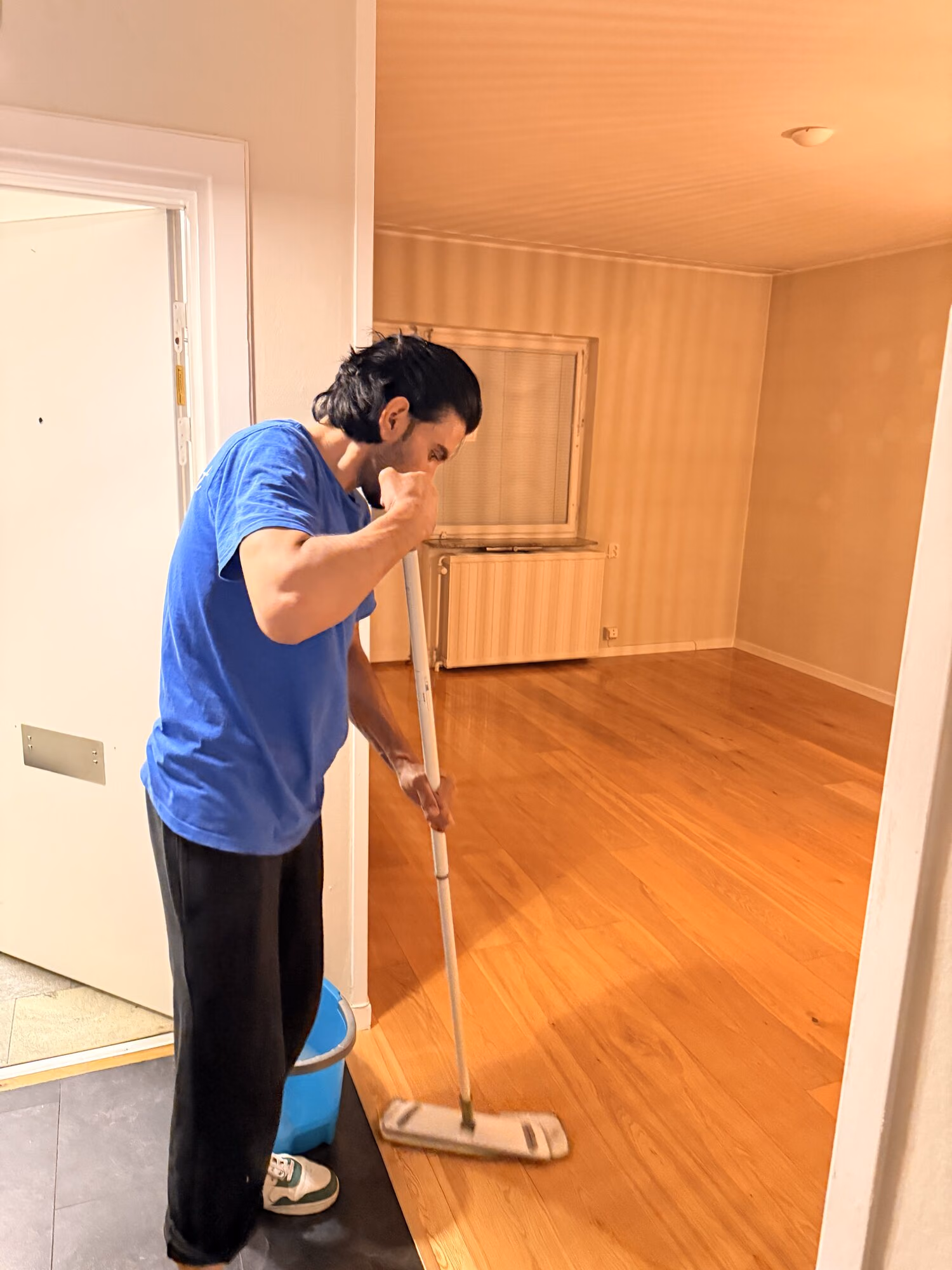Städare moppar ett parkettgolv i en tömd lägenhet med en blå hink bredvid sig