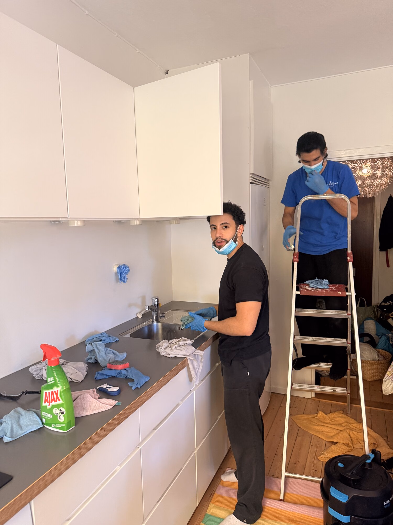 Två städare i handskar och munskydd rengör ett kök med trappstege, mikrofiberdukar och dammsugare