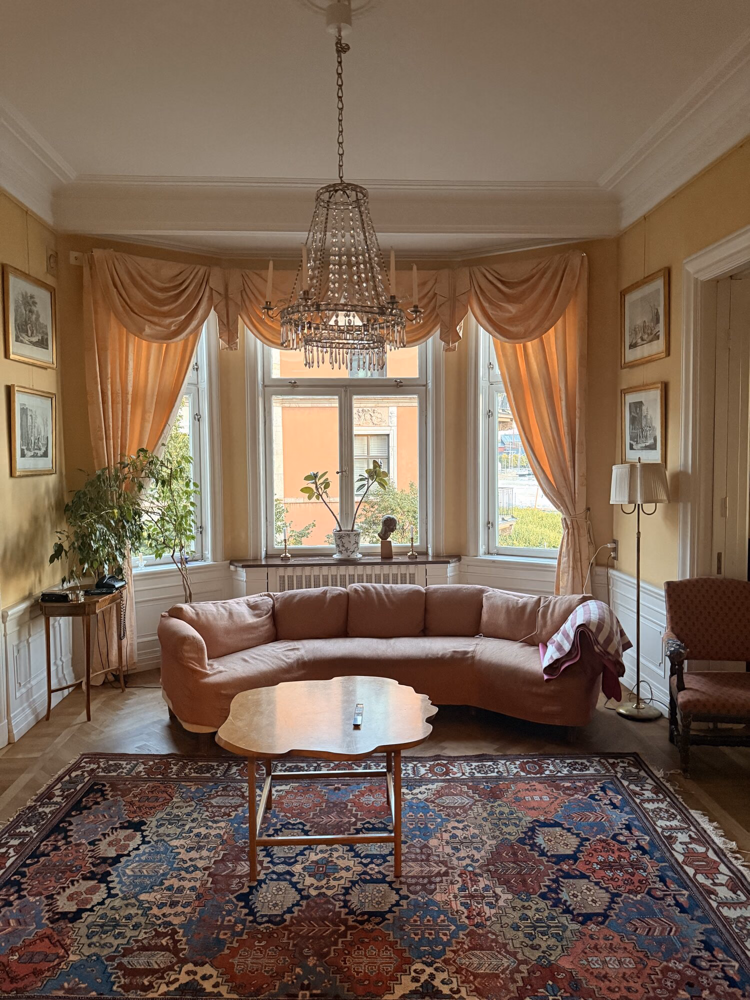 Ljust sekelskiftesvardagsrum med kristallkrona, rosa soffa och stor orientalisk matta på fiskbensparkett